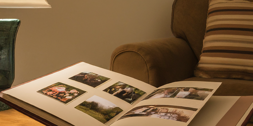 An open photo album on a wooden table under the warm light of a lamp, beside a cozy armchair with striped cushions.
